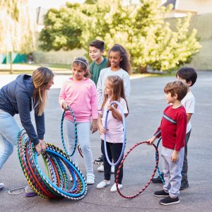 Children in summer camp with sports teacher Eine Frau verteilt bunte Hula-Hoop-Reifen an sechs Kinder in einem sonnigen Außenbereich der Danielschule. Die leger gekleideten Kinder stehen in einem Halbkreis, aufgeregt und engagiert. Im Hintergrund sind Bäume und ein Gebäude zu sehen, die dieser lebendigen Szene des spielerischen Lernens Wärme verleihen.