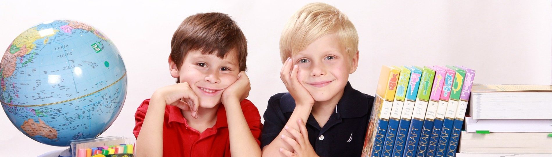 boys-286245_1920 Zwei Jungen sitzen an einem Tisch, umgeben von aufgeschlagenen Büchern, einem Stapel Enzyklopädien und einer Schachtel Buntstifte, daneben ein Globus. In ihrer christlichen Gesamtschule trägt der eine ein rotes Hemd, der andere ein marineblaues; beide lächeln strahlend und stützen ihre Hände auf das Gesicht.