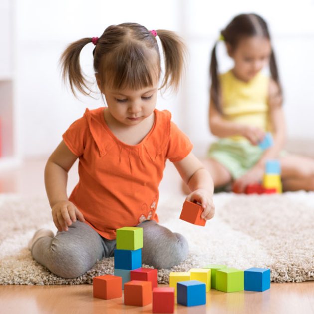 Zwei kleine Kinder sitzen auf einem Teppich in der Danielschule und spielen vertieft mit bunten Bauklötzen. Das Kind im Vordergrund trägt ein orangefarbenes Hemd und eine graue Hose und ist auf das Stapeln von Bauklötzen konzentriert, während das Kind in Gelb im Hintergrund ebenfalls sehr beschäftigt ist.