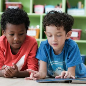 Zwei Jungen liegen auf dem Boden eines Klassenzimmers in einer christlichen Gesamtschule und teilen sich ein Buch. Der eine trägt ein rotes Hemd, der andere ein blaues. Hinter ihnen stehen grüne Regale mit bunten Behältern. Sie sind eifrig in ihr gemeinsames Leseabenteuer vertieft.