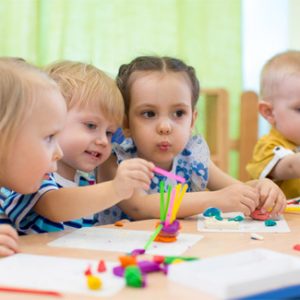 Vier kleine Kinder sitzen um einen Tisch in der Danielschule und beschäftigen sich mit einem Kunstprojekt. Sie formen bunte Knete zu Formen und Figuren. Der Raum ist hell, ein sanfter grüner Vorhang bildet einen ruhigen Hintergrund und spiegelt die fürsorgliche Umgebung dieser christlichen Gesamtschule wider.