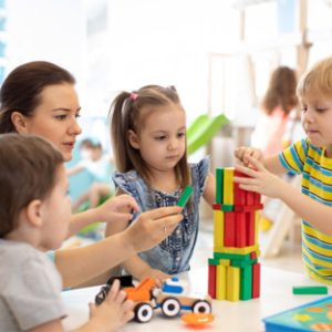 In einem hellen Raum in der christlichen Gesamtschule spielen eine Frau und drei kleine Kinder fröhlich mit bunten Bauklötzen am Tisch. Ein Kind stapelt Klötze, während die anderen zuschauen und weitere Klötze halten. In der Nähe steht ein Spielzeuglastwagen, der die spielerische Szene noch verstärkt.