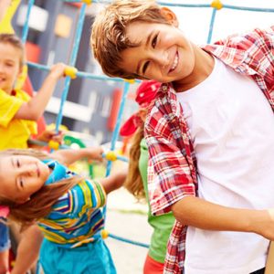 faq-01 Kinder spielen auf den bunten Spielgeräten der Christlichen Schule. Ein Junge in einem rotkarierten Hemd und einem weißen T-Shirt lächelt in die Kamera und verkörpert die Freude und Kameradschaft dieser lebendigen Gemeinschaft. Andere Kinder in heller Kleidung klettern und haben Spaß im Hintergrund.