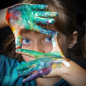 gallery3 Ein Kind aus einer christlichen Gesamtschule hält seine Hände hoch, um sein Gesicht zu umrahmen, die Handflächen sind mit leuchtender, bunter Farbe bedeckt. Die blauen Augen blicken durch den Handrahmen. Der Hintergrund ist dunkel und hebt die bunte Farbe und den konzentrierten Ausdruck des Kindes hervor.