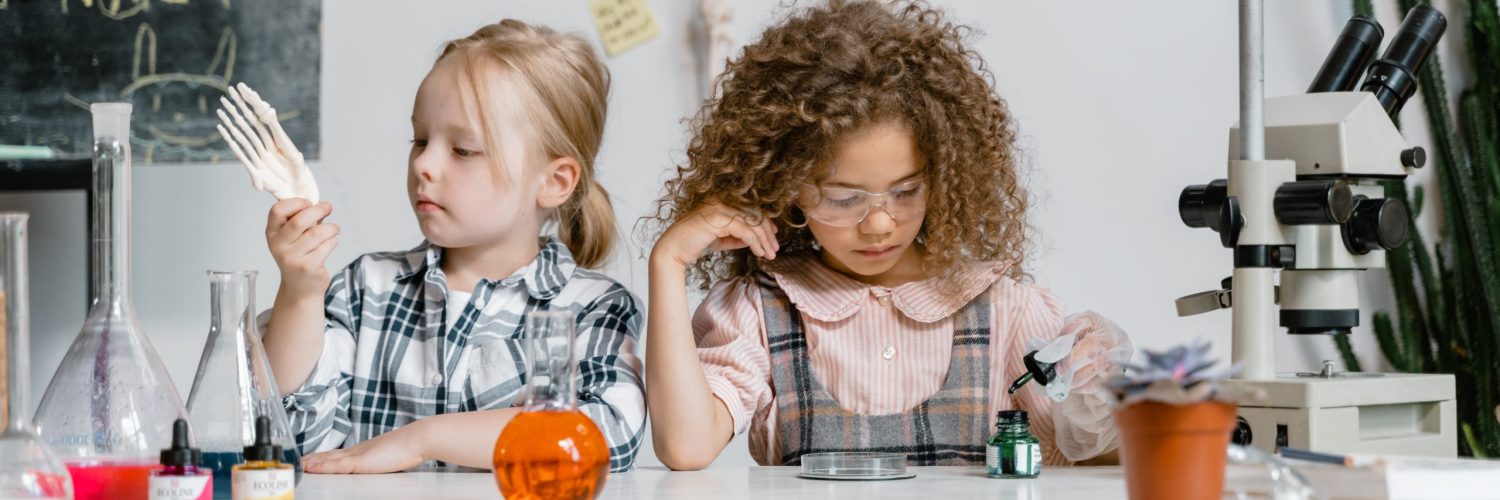 pexels-mart-production-8471975 In einem lebendigen naturwissenschaftlichen Klassenzimmer der Danielschule erleben zwei Kinder ein Lernabenteuer. Das eine hantiert mit Plastikutensilien, während das andere unter eine Lupe blickt. Naturwissenschaftliche Geräte und eine Pflanze füllen den Tisch, ein Mikroskop und eine Kreidetafel deuten auf weitere Entdeckungen dahinter hin.
