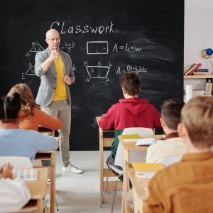 pexels-max-fischer-5212345 In einem Klassenzimmer einer christlichen Gesamtschule steht ein Lehrer vor einer Tafel mit geometrischen Diagrammen und dem Wort „Klassenarbeit“ und unterrichtet aufmerksame Schüler. Ein Schüler im roten Hemd hebt die Hand, während andere an Holzpulten sitzend zusehen.